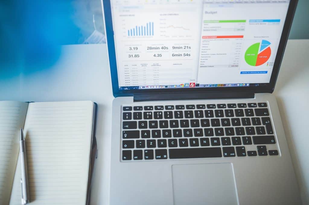 Laptop on a desk showing charts and a budget table, next to an open notebook and pen